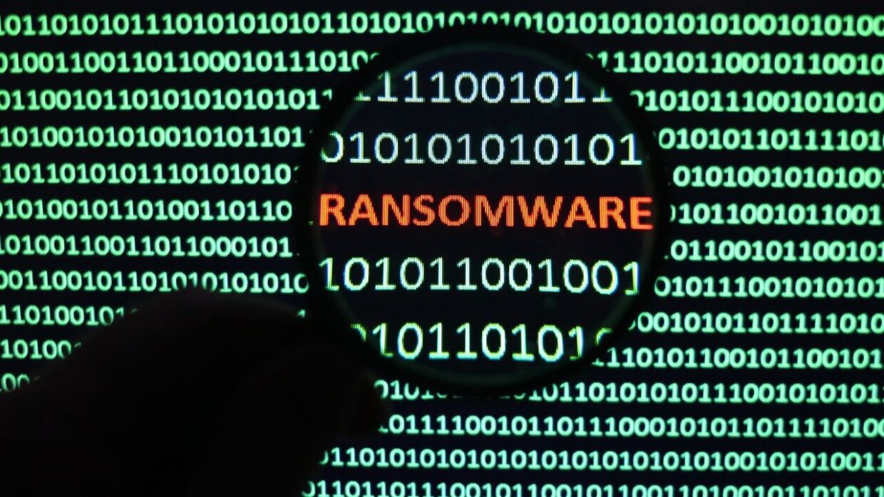 A magnifying glass overlaid on a screen displaying binary code, highlighting the word 'RANSOMWARE' in orange amidst green background.