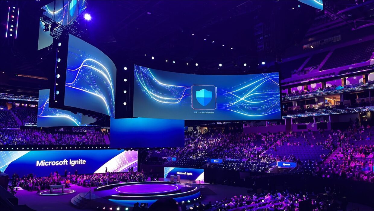A large auditorium filled with audience members attending the Microsoft Ignite event. The stage features multiple large screens displaying vibrant blue graphics, with the Microsoft Ignite logo in focus.