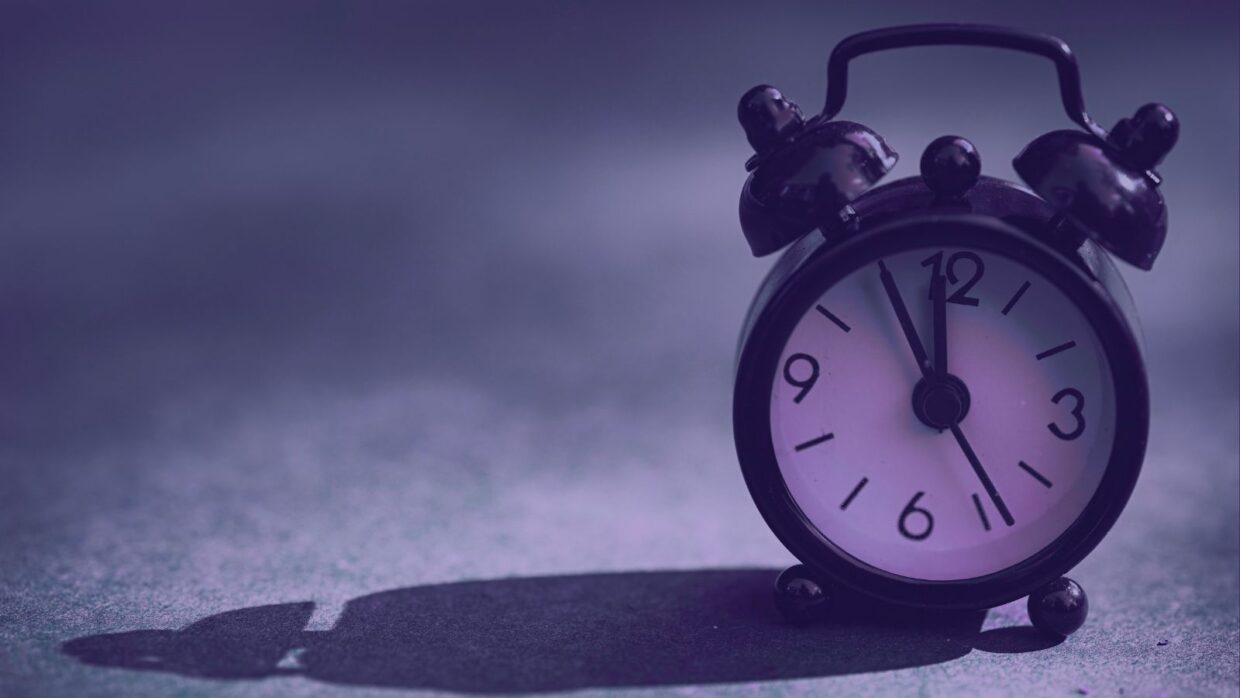 A vintage black alarm clock showing 12:00, positioned on a textured surface with a purple tint. The shadow of the clock is visible on the surface.