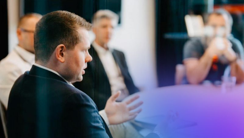 A professional man speaking during a meeting, gesturing with his hand, while a small group of colleagues listen attentively in a modern conference room.