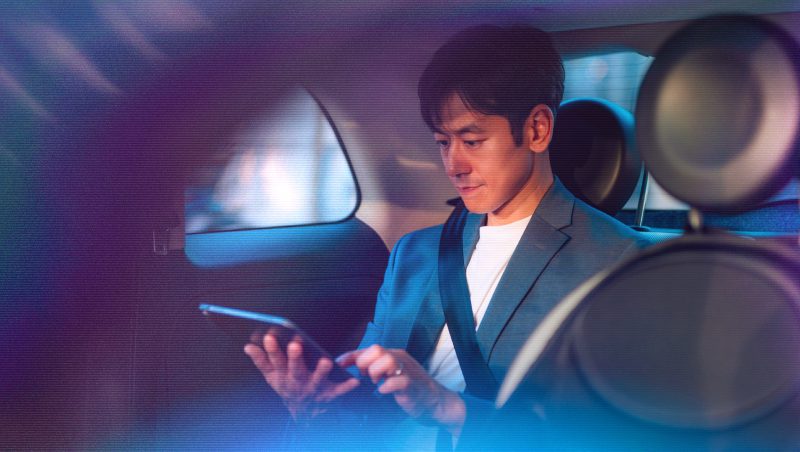 A man dressed in a suit sitting in the back seat of a car, holding a smartphone and looking at the screen. The interior of the car is softly lit.