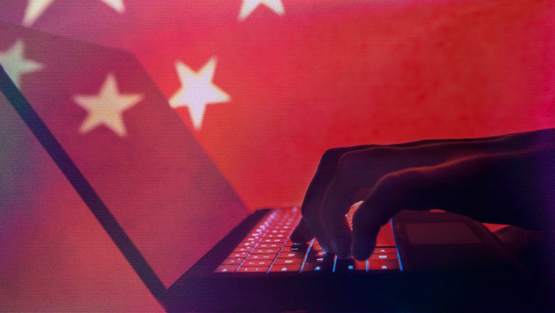A silhouetted hand typing on a laptop with illuminated keys, set against a backdrop of a red surface featuring white stars, resembling the flag of China.
