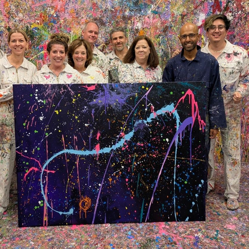 A group of colleagues smiling and waving during a celebration, wearing gray jackets, in front of a dark blue backdrop. A 'Great Place To Work' certification badge is displayed in the corner.
