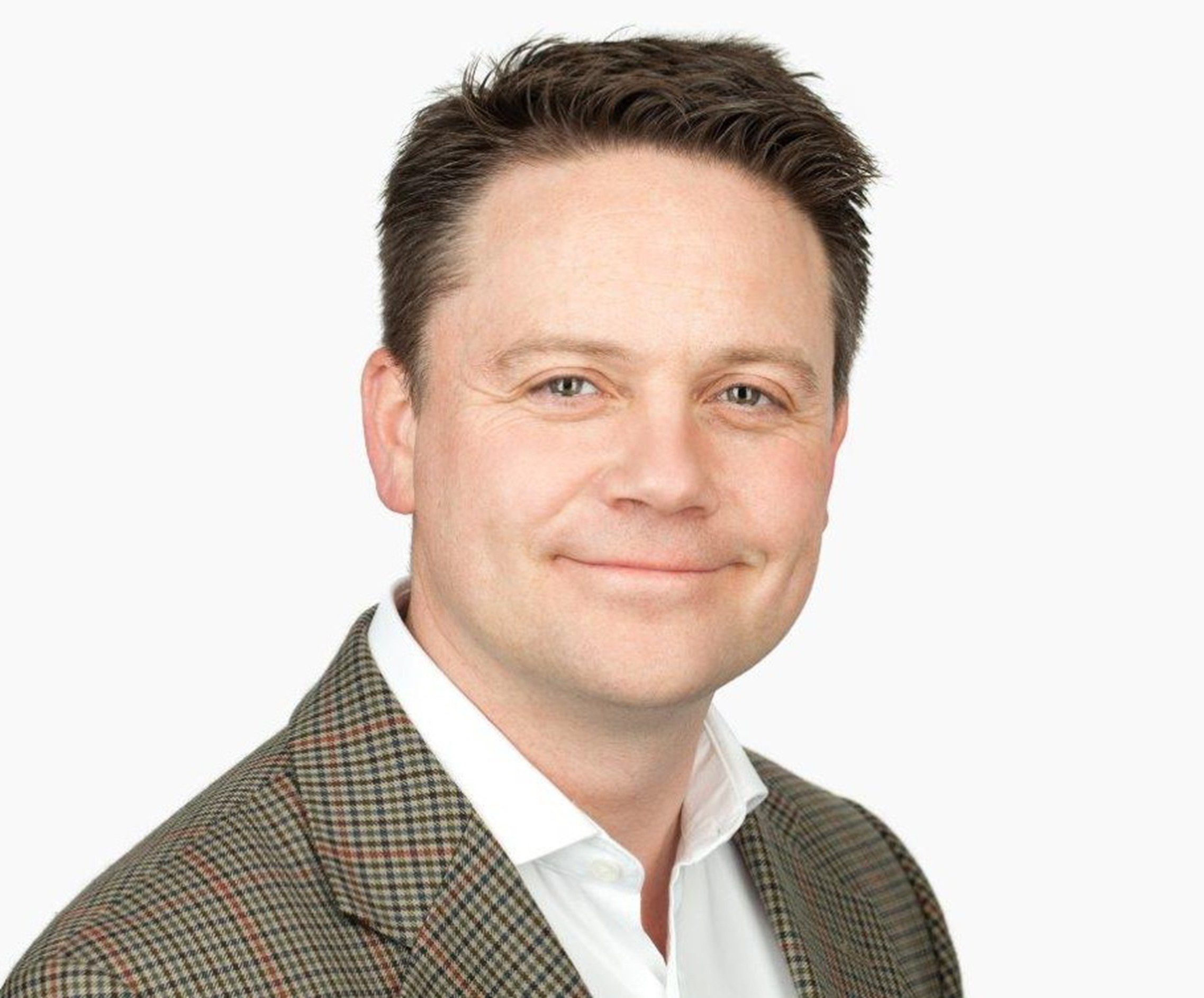 A professional headshot of a man smiling. He has short dark hair and is wearing a brown checkered blazer over a white shirt. The background is plain white, providing a clean and clear view of his face.