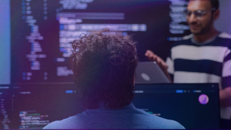 A person with curly hair is seated at a computer, facing a monitor displaying coding lines. In the background, another person is gesturing while speaking, wearing a striped shirt. The setting appears to be a tech-focused work environment.