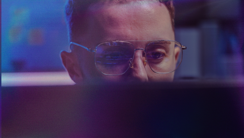 A man wearing glasses looks at a screen in a dimly lit room with blue and purple lighting.