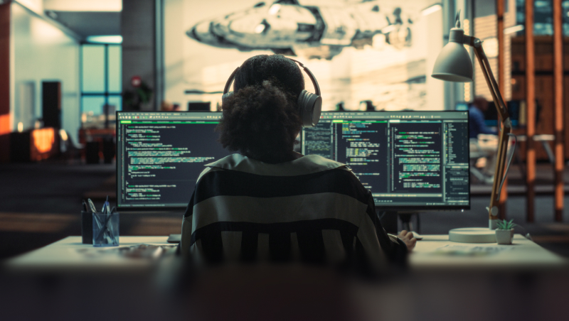 A person wearing headphones sits at a desk facing dual computer monitors with code on screen, in a modern office workspace.